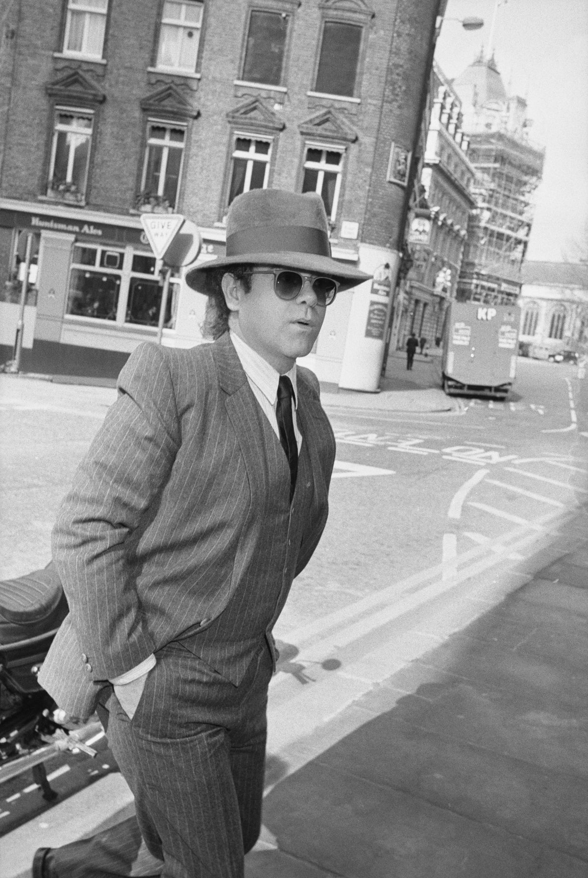 Black and white photograph of Elton John in a pinstripe three-piece suit, fedora and sunglasses walking down a London street, 1985