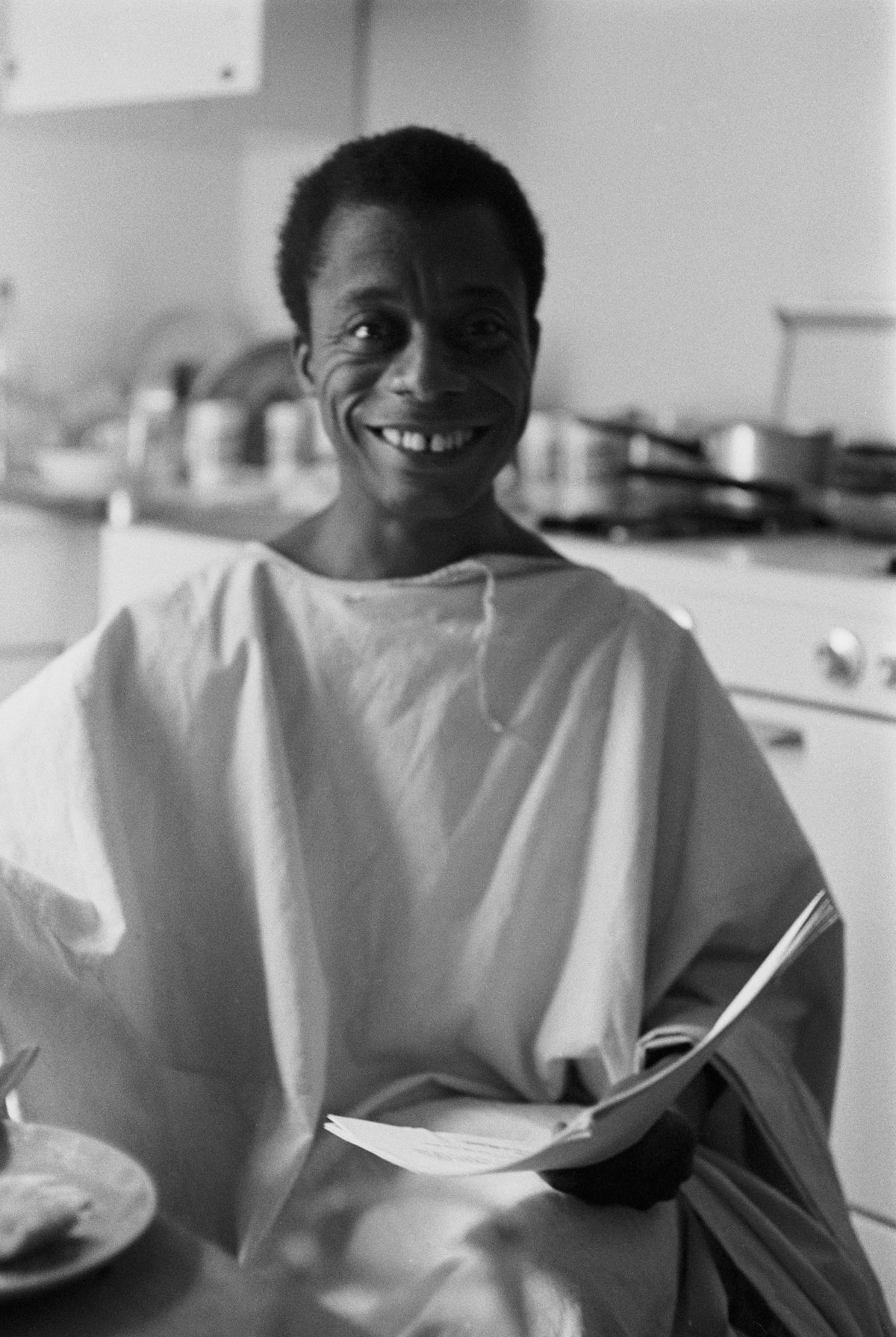 Black and white photograph of a smiling James Baldwin seated in a kitchen holding a sheaf of papers, USA, 1963