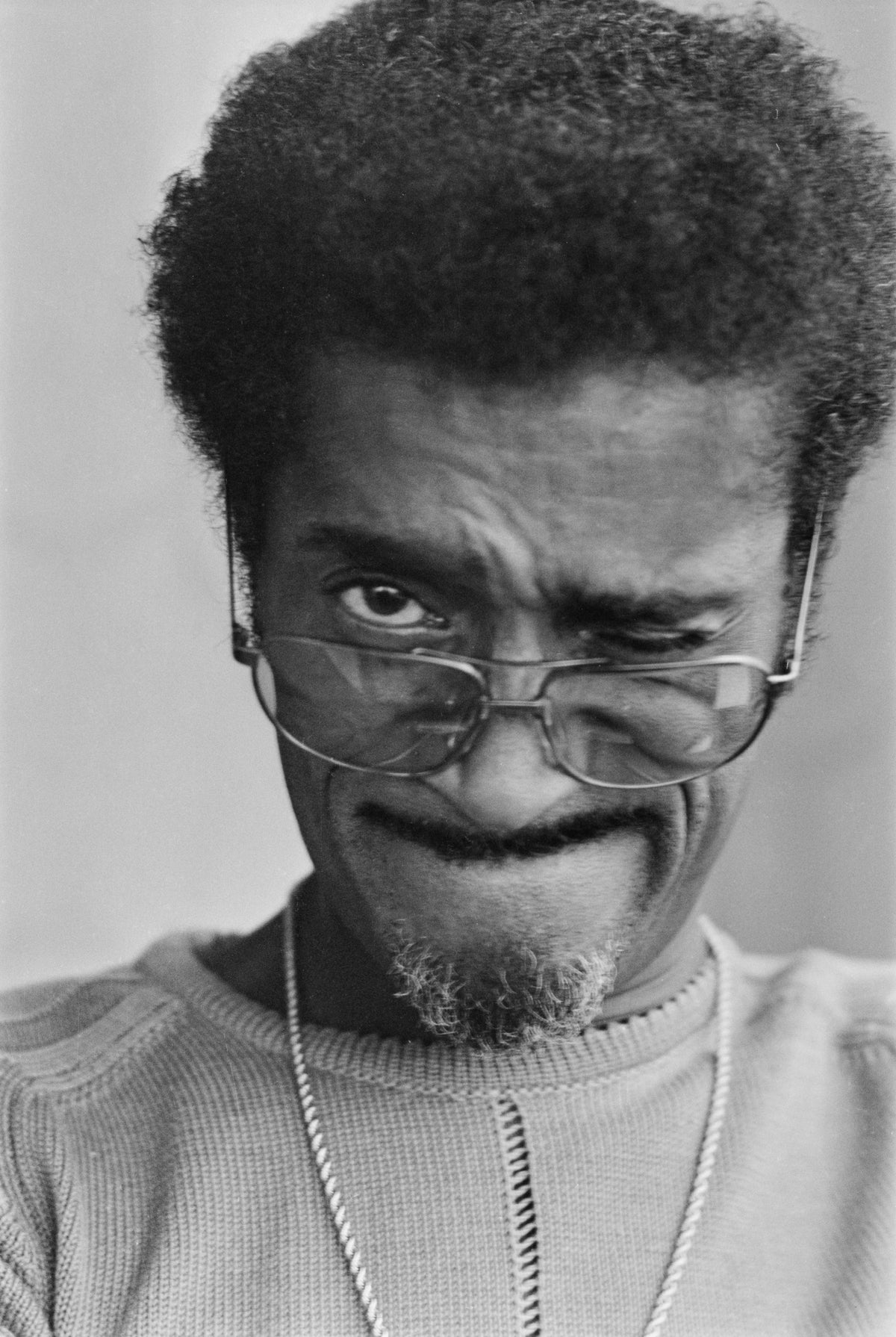 Black and white close-up of Sammy Davis Jr pulling a comic face, squinting one eye behind his glasses, London, 1972