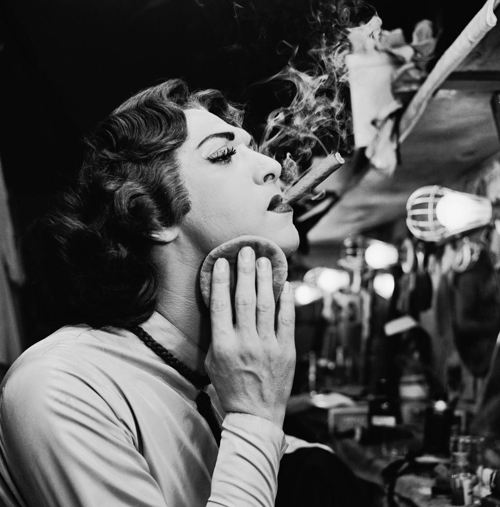 Black and white photograph of a female impersonator powdering their face with a cigarette in hand, backstage at a mirror, circa 1955