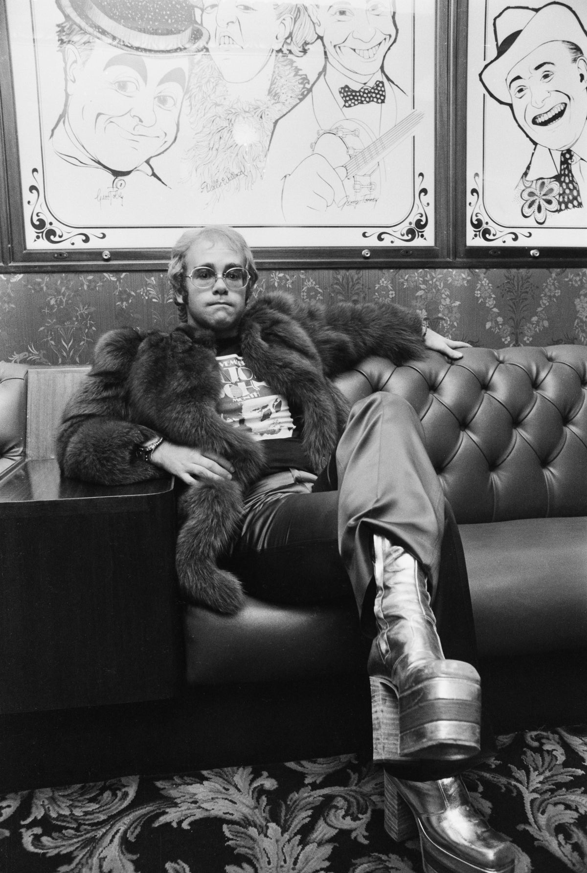 Black and white photograph of Elton John in a fur coat and platform boots lounging on a chesterfield in the London Palladium bar, 1972
