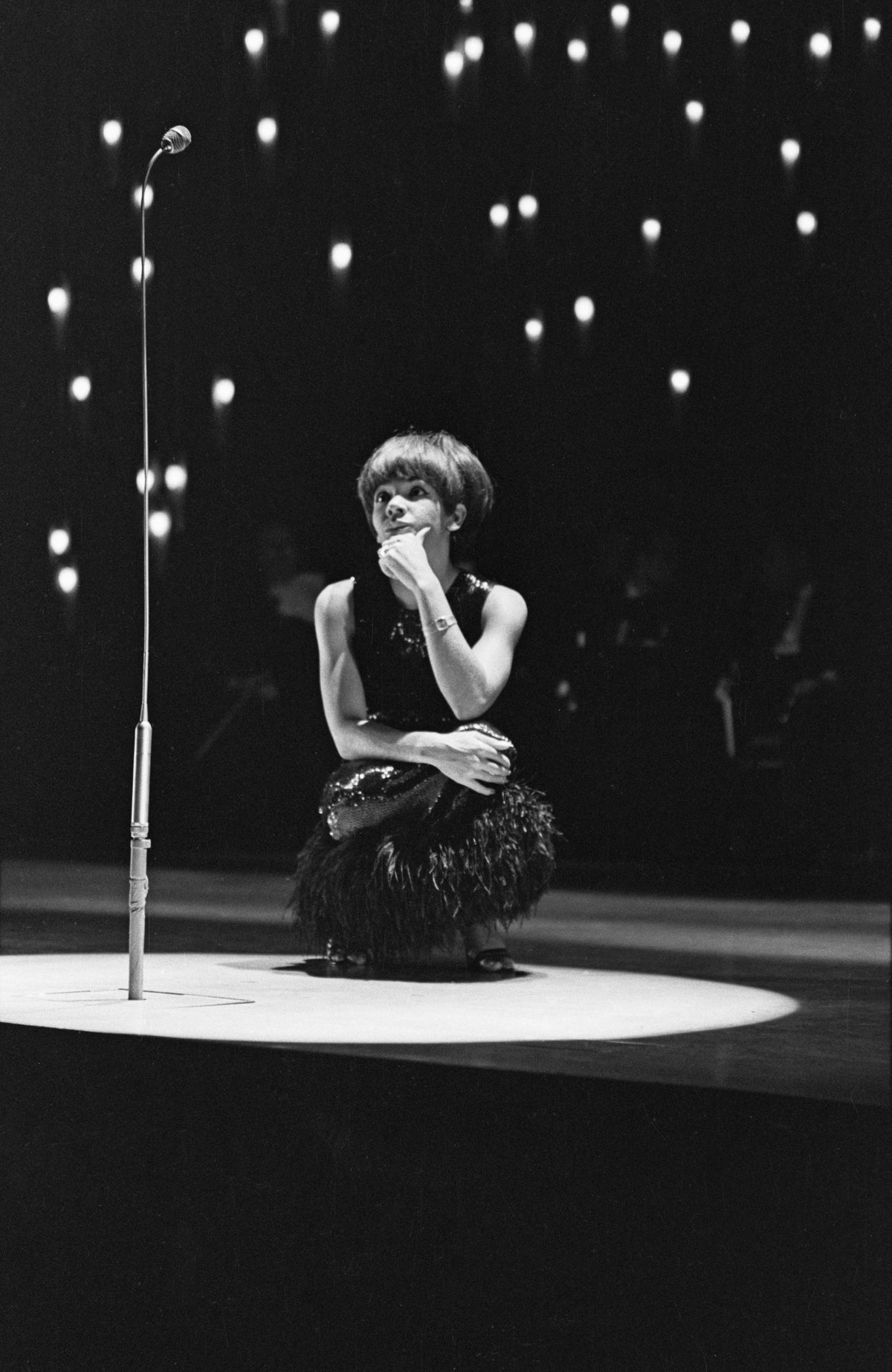 Black and white photograph of Shirley Bassey crouching on a spotlit stage beside a microphone stand at the London Palladium, 1965