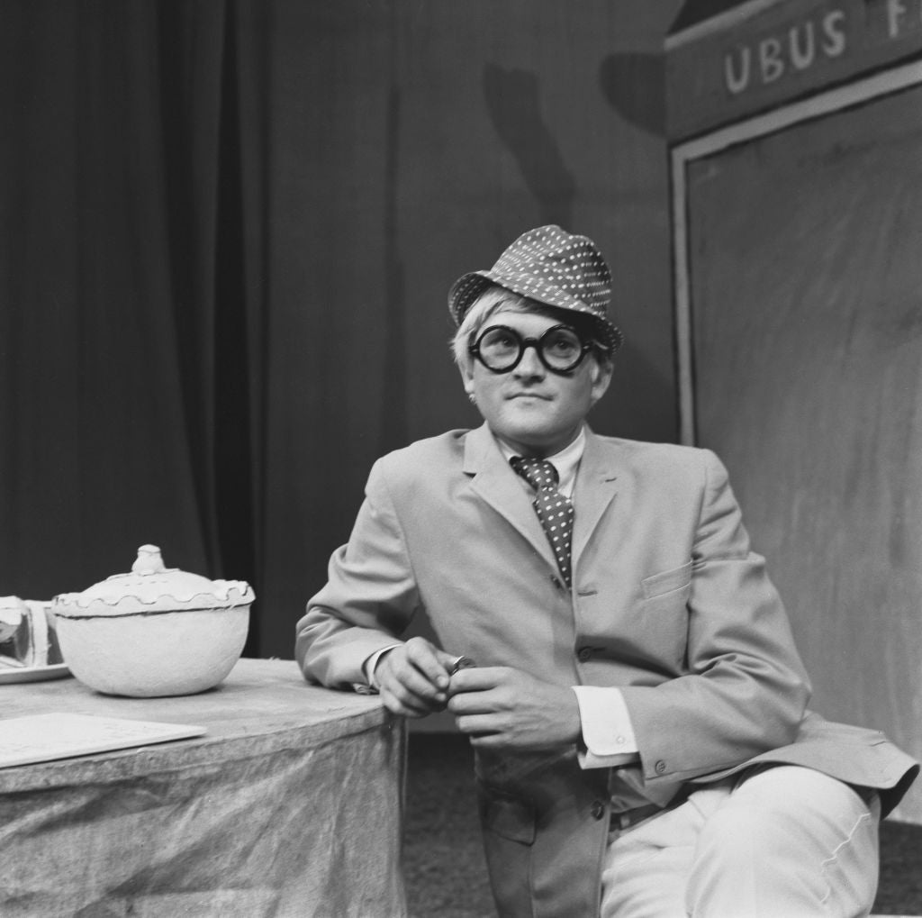 Black and white photograph of David Hockney in a hat, round glasses and suit on the set of Ubu Roi at the Royal Court Theatre, 1966