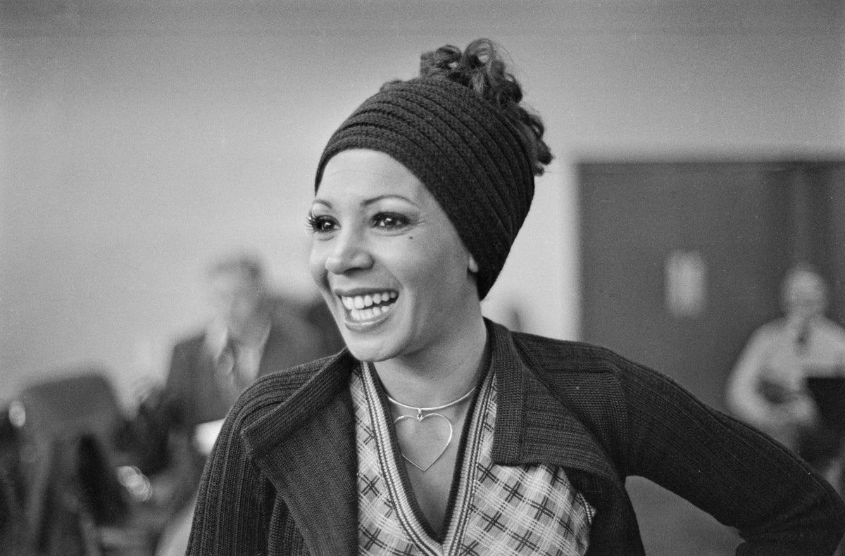 Black and white photograph of a smiling Shirley Bassey in a knitted headband and checked top, UK, 1973
