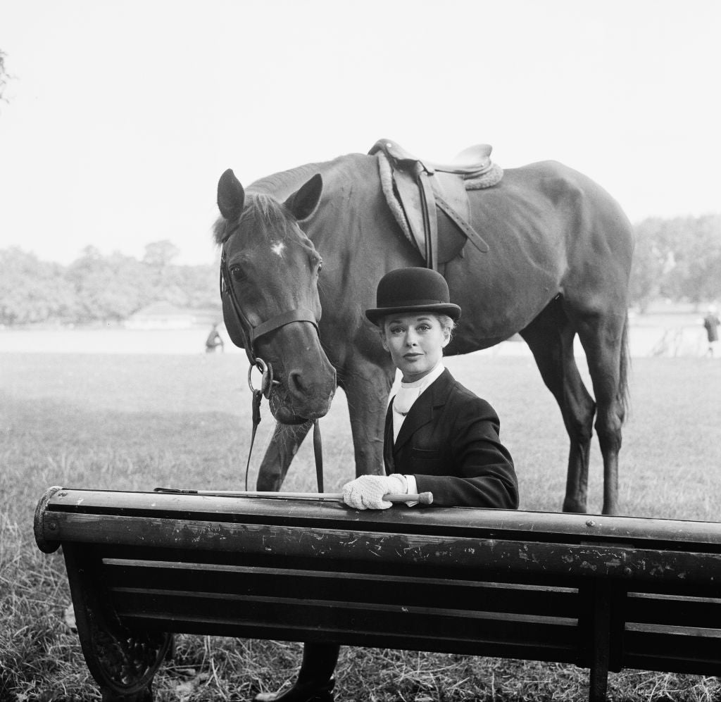 Black and white photograph of Tippi Hedren in a bowler hat beside a saddled horse behind a park bench, London, 1963