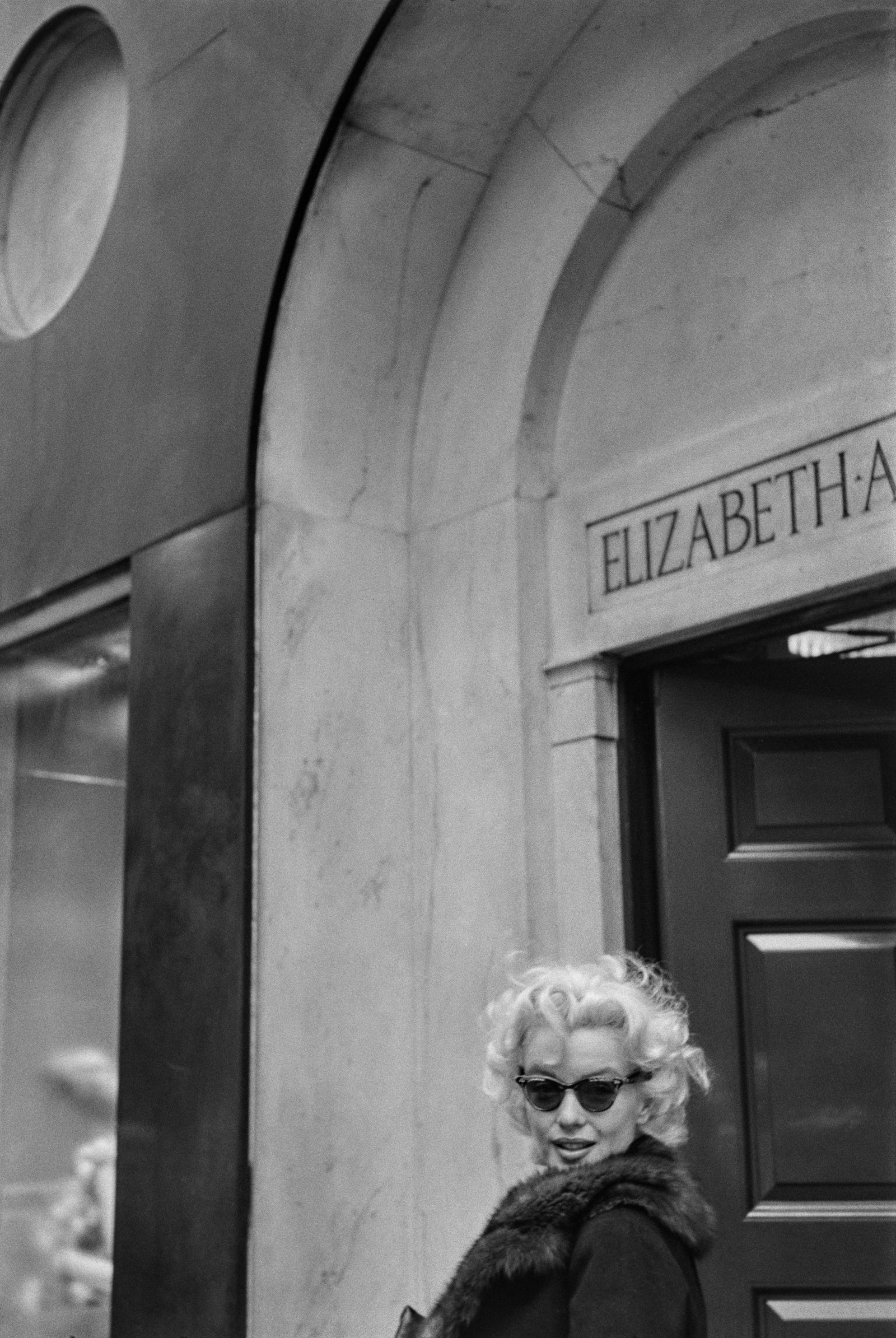 Black and white photograph of Marilyn Monroe in sunglasses outside the Elizabeth Arden salon, New York, 1955