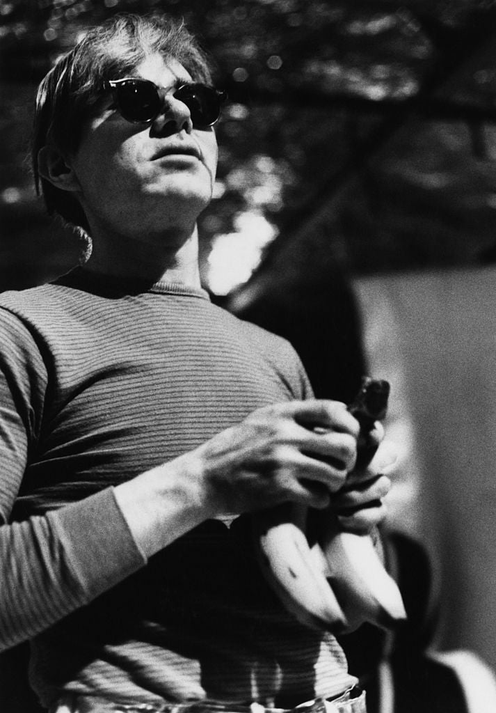 Black and white photograph of Andy Warhol in dark sunglasses and a striped top holding a bunch of bananas, circa 1965