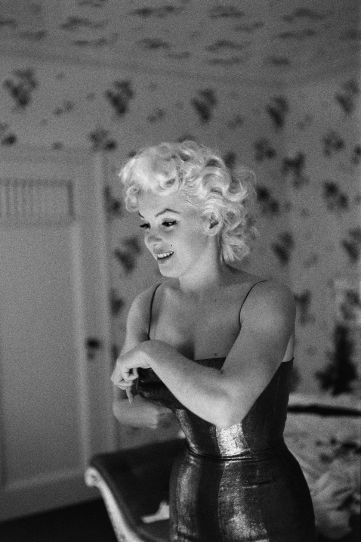 Black and white photograph of Marilyn Monroe adjusting her strapless dress in a floral-wallpapered room at the Ambassador Hotel, New York, 1955
