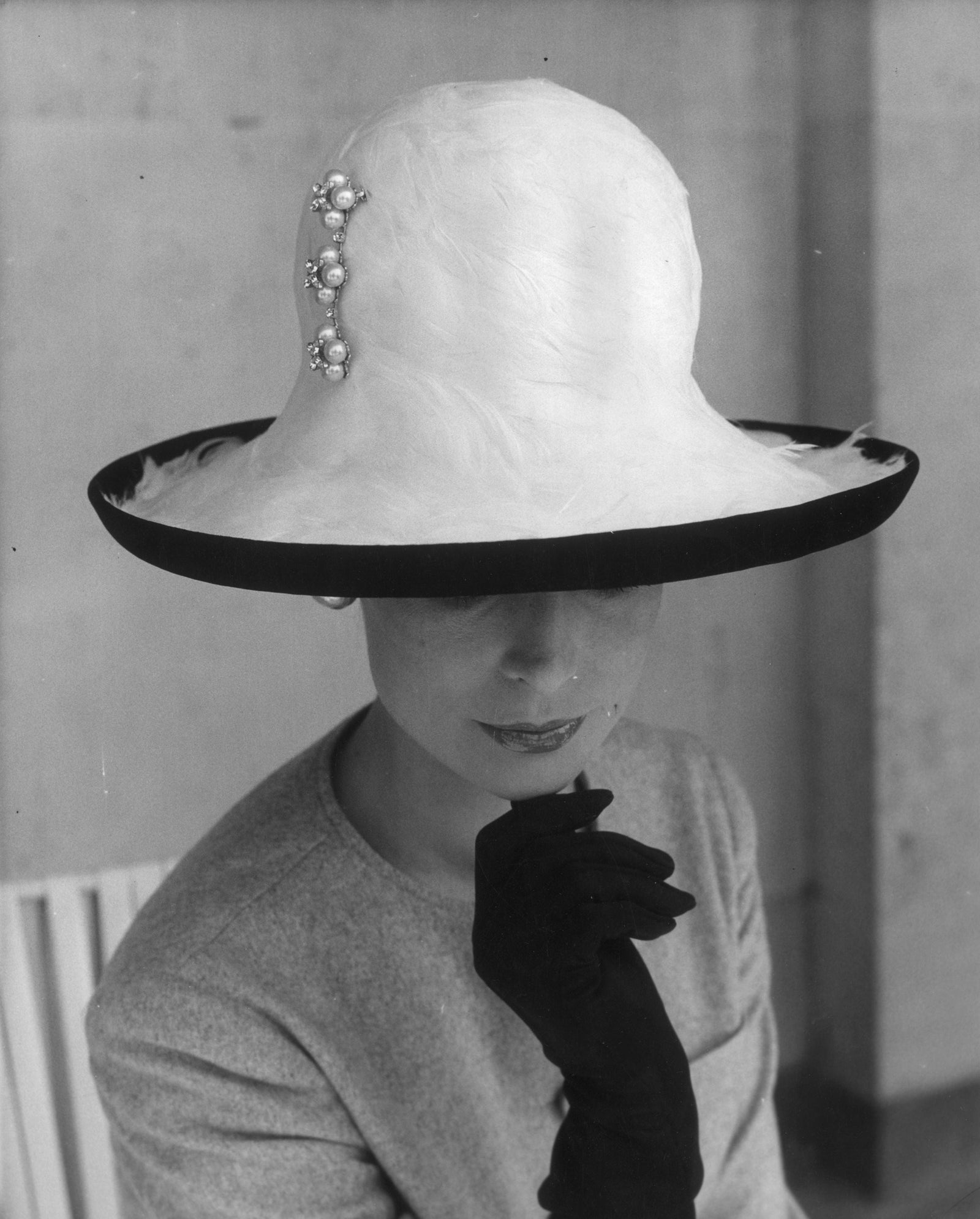 Black and white photograph of model Brenda Harper in a wide-brimmed jewelled hat, gloved hand raised to her chin, London, 1964