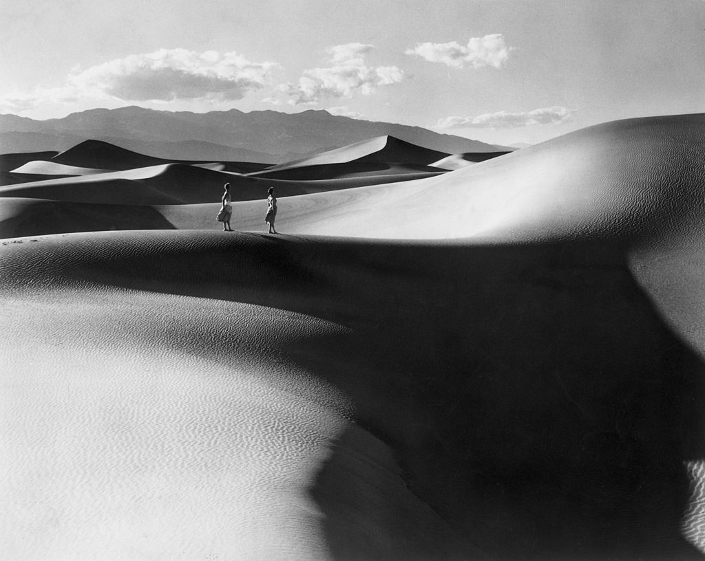 Black and white photograph of two tiny figures walking across the sweeping sand dunes of Death Valley