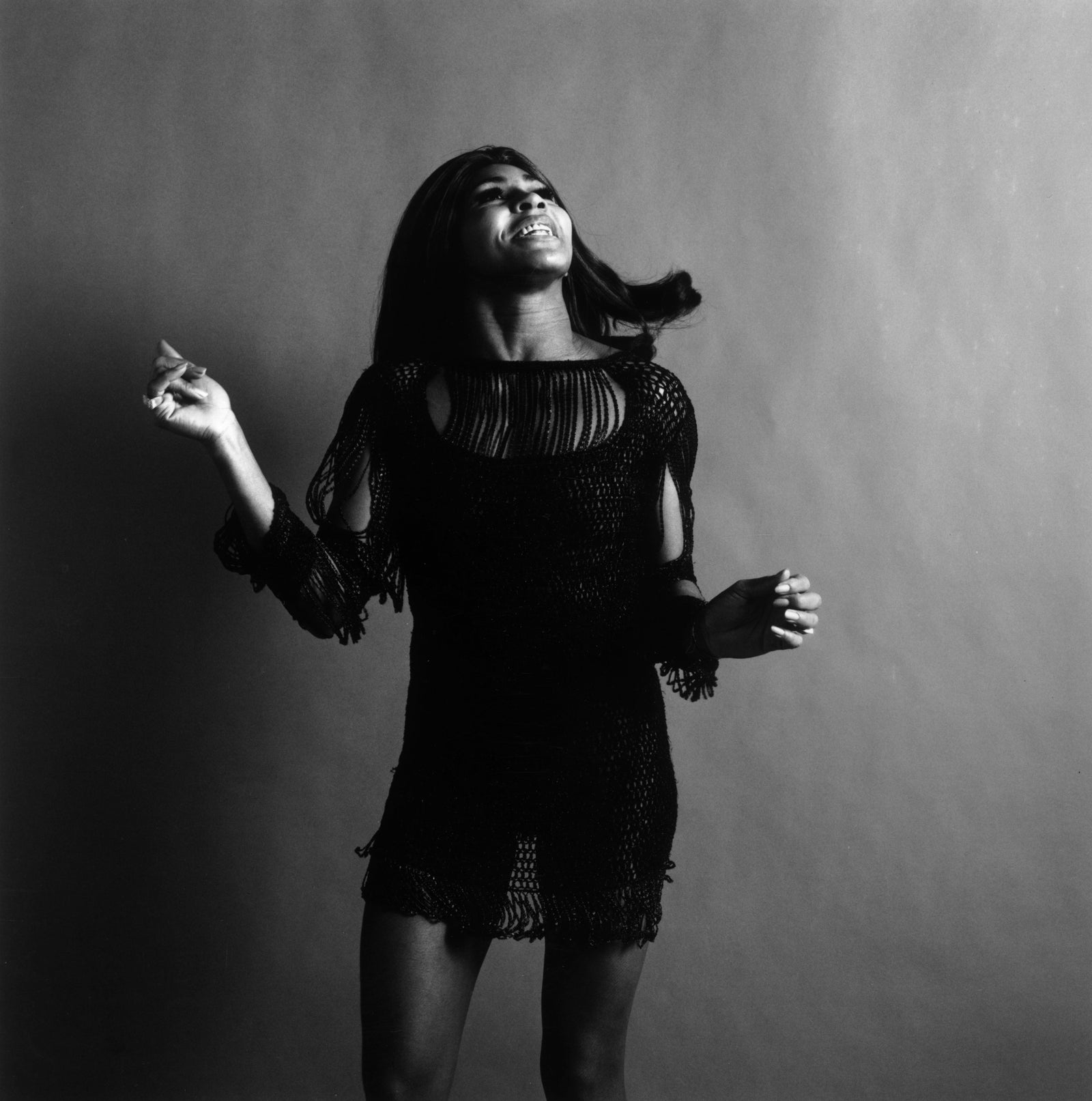 Black and white studio portrait of Tina Turner in a dark crochet mini dress, looking up and snapping her fingers, 1969