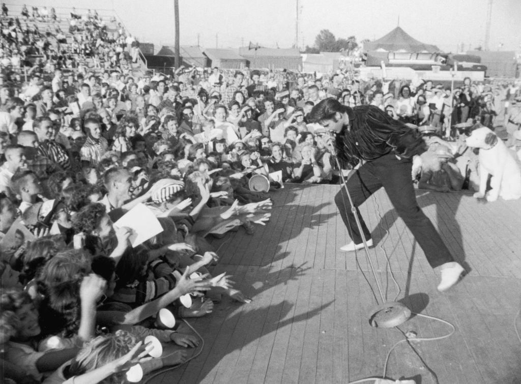 Black and white photograph of Elvis Presley performing on a small outdoor stage in front of a large crowd of young fans, circa 1957