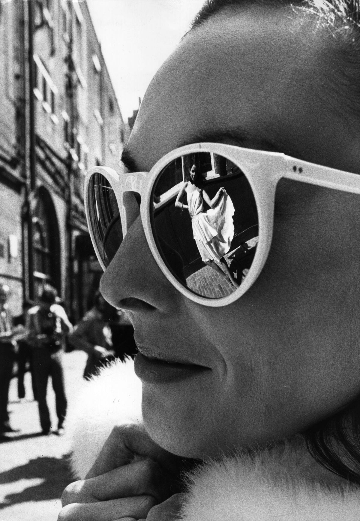 Black and white close-up of a woman in round white sunglasses with a street scene reflected in the lenses, 1980