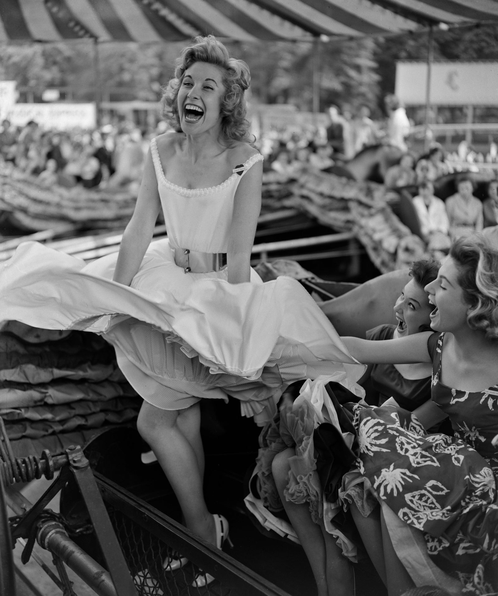 Black and white photograph of three laughing women with skirts billowing in the wind machine at the Battersea Park fun fair, London, 1957