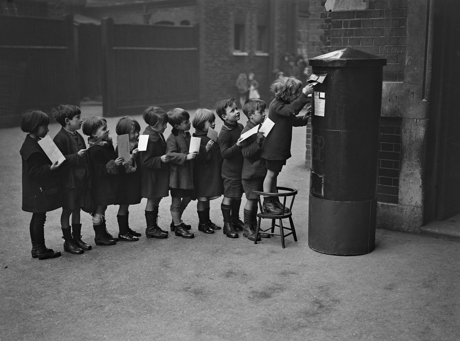 Black and white photograph of a queue of schoolchildren posting letters into a pillar box in a London school playground, 1926