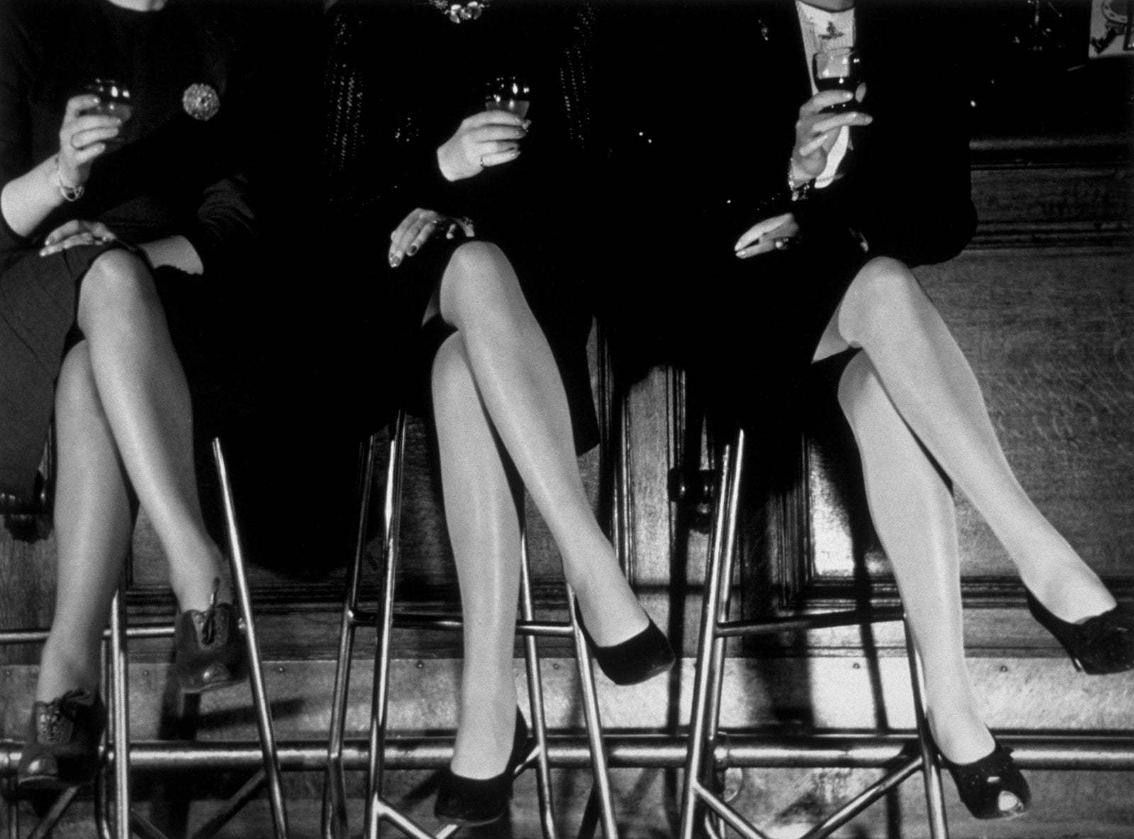 Black and white photograph of three pairs of crossed stockinged legs in high heels on bar stools, 1946