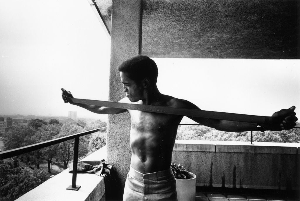 Black and white photograph of Sammy Davis Jr stretching a chest expander on a concrete balcony, 1968