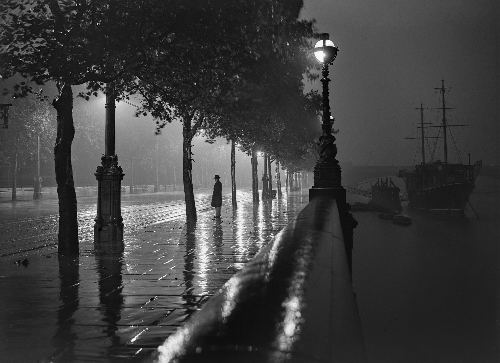 Black and white photograph of a lone figure on the rain-soaked Thames Embankment at night under street lamps, London, 1929