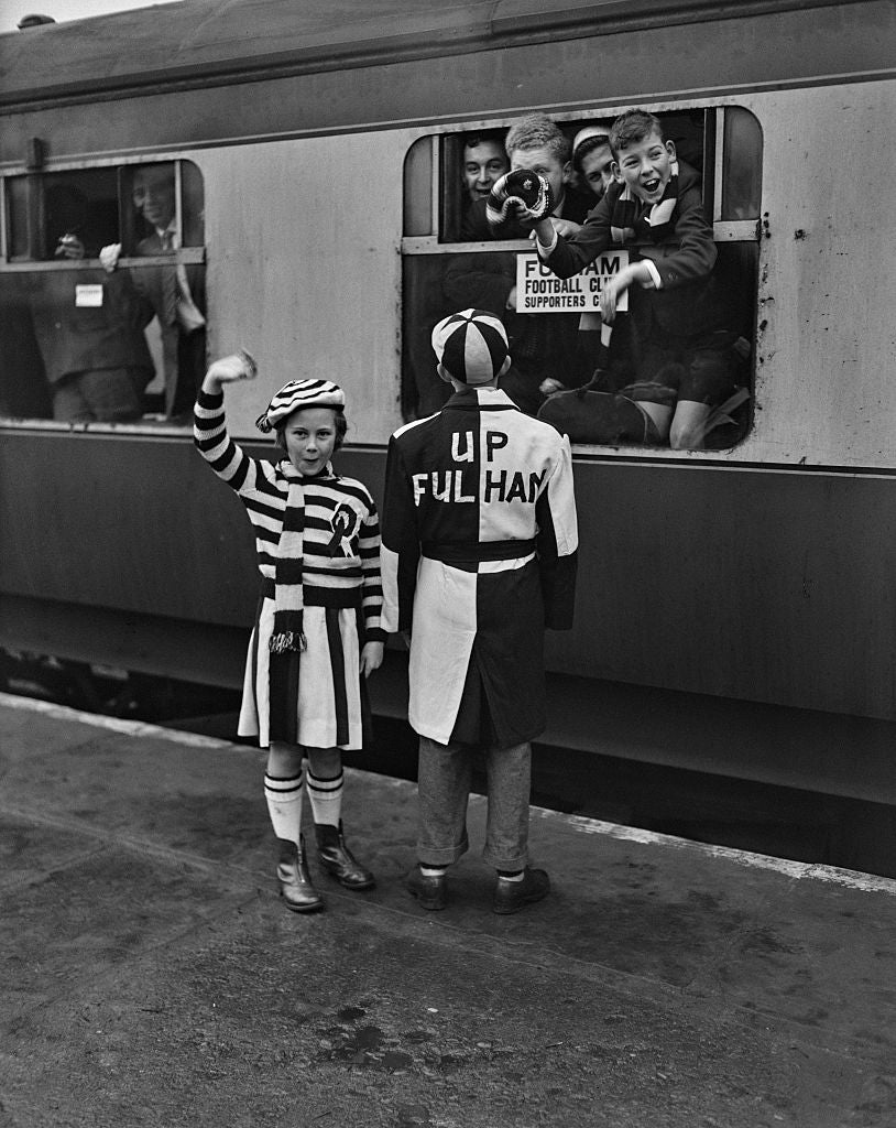 Black and white photograph of two young Fulham fans in black and white outfits waving at a football special train, London, 1957