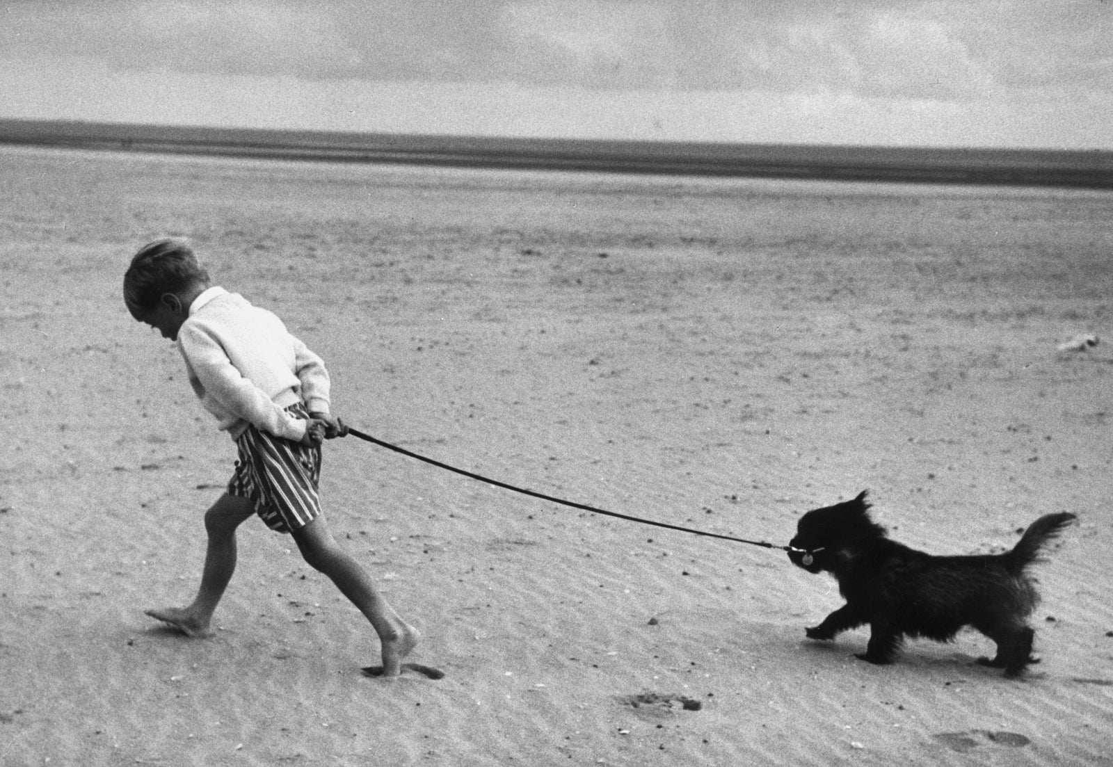 Black and white photograph of a barefoot young boy walking his small black dog on a lead across Camber Sands, 1959