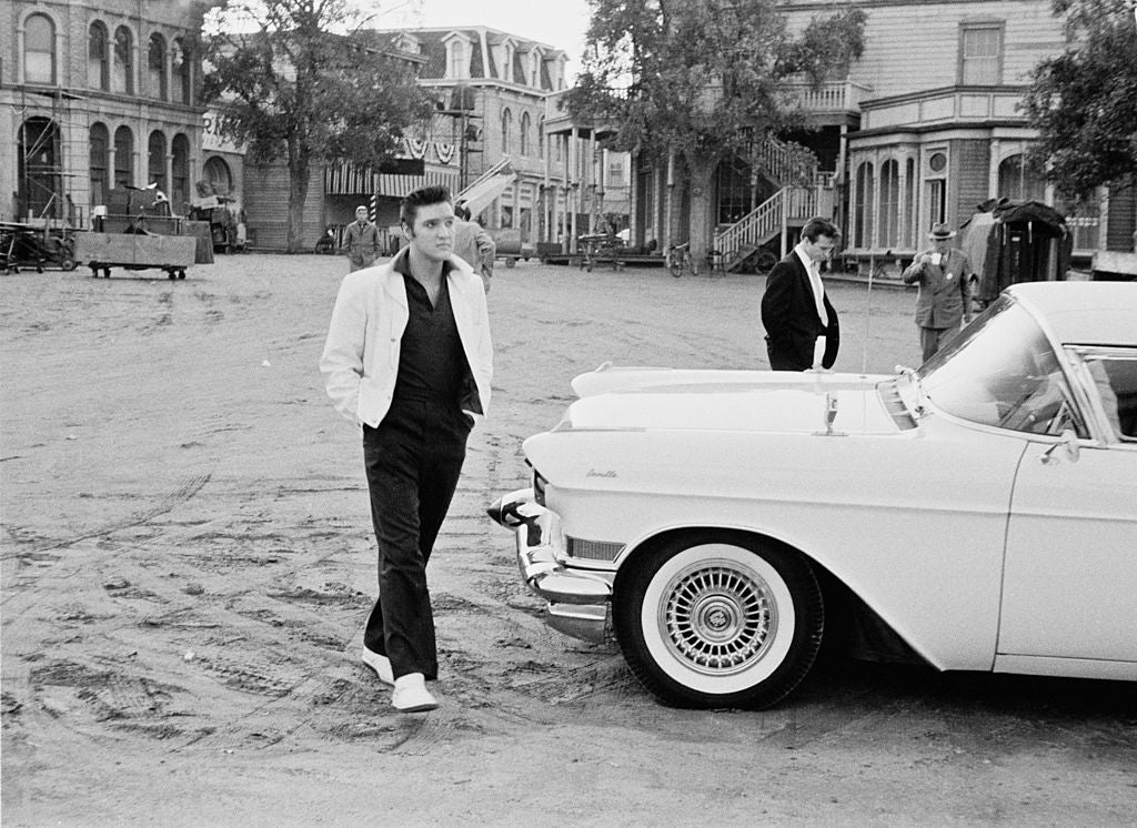 Black and white photograph of Elvis Presley walking past a white Cadillac Eldorado on a film set, circa 1958