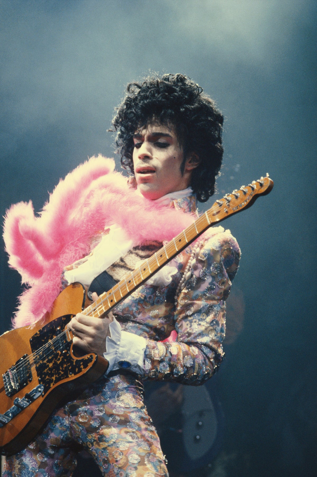 Colour photograph of Prince playing guitar on stage in a paisley suit with a pink feather boa at the Forum, Inglewood, 1985