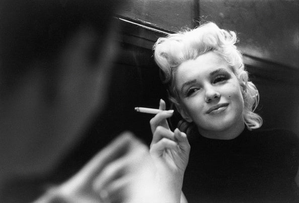 Black and white photograph of Marilyn Monroe smiling softly with a cigarette in a dimly lit New York restaurant, 1955