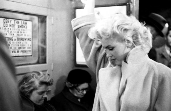 Black and white photograph of Marilyn Monroe in a pale coat looking down inside a subway carriage, New York, 1955