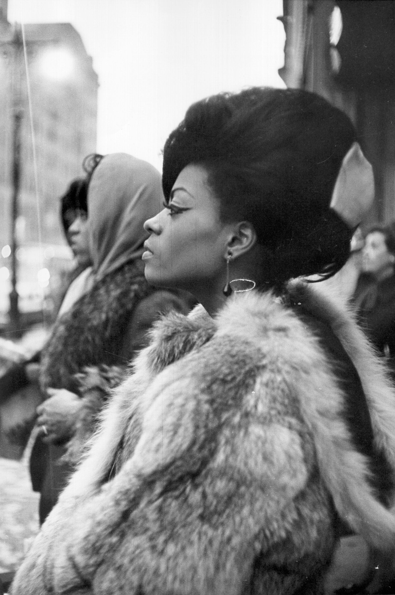 Black and white photograph of a member of The Supremes in profile wearing a fur coat and bouffant hairstyle on a city street