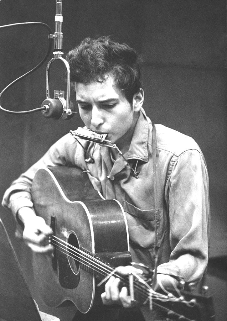 Black and white photograph of Bob Dylan playing acoustic guitar and harmonica at a microphone while recording his first album, New York, 1961