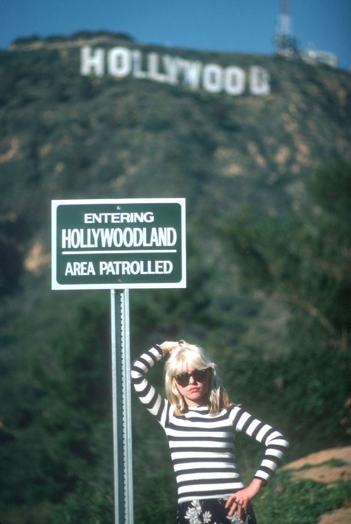 Colour photograph of Debbie Harry of Blondie in a striped top beside an Entering Hollywoodland sign beneath the Hollywood Sign, 1977