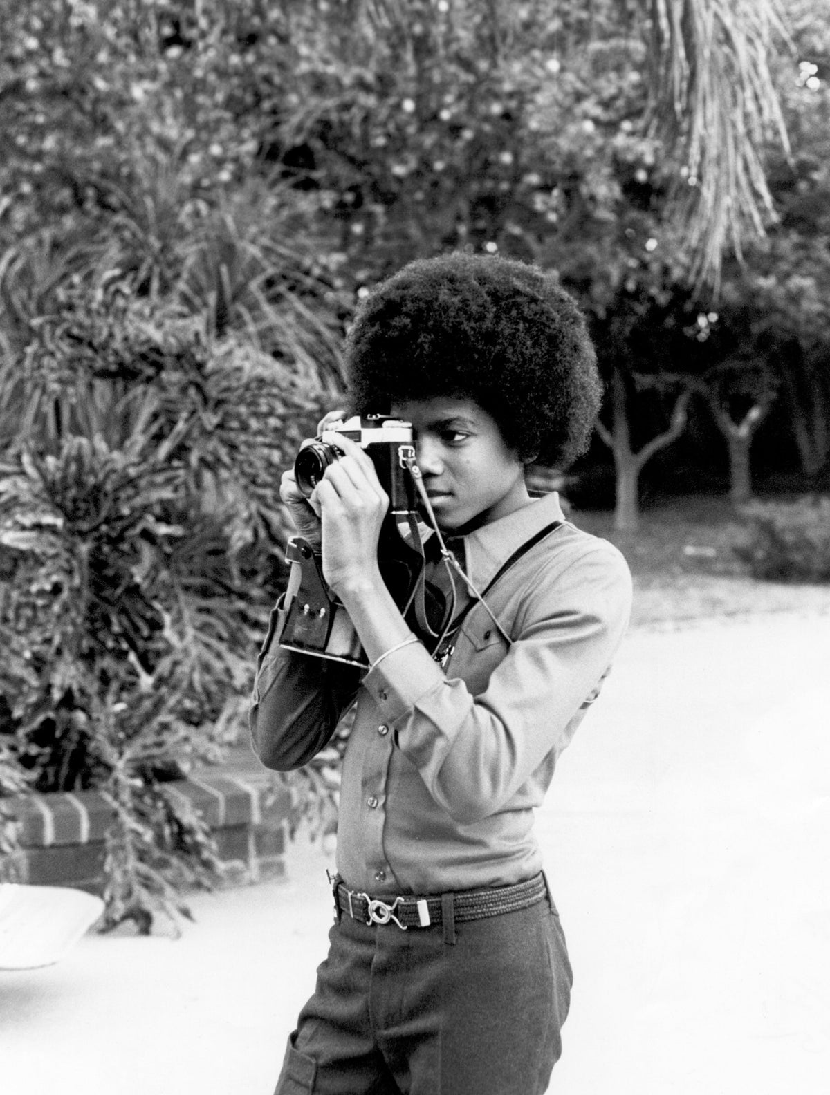 Black and white photograph of a young Michael Jackson taking a picture with a camera in the garden of his Hollywood Hills home, 1972