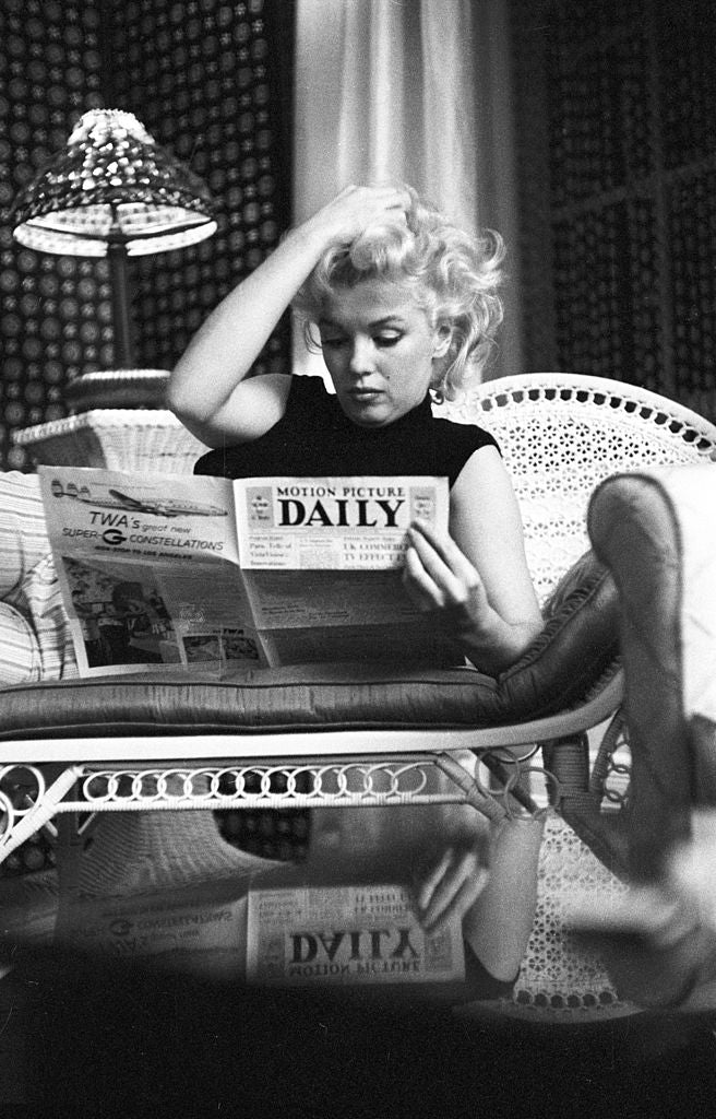 Black and white photograph of Marilyn Monroe reading Motion Picture Daily on a wicker chaise beneath a lamp, Ambassador Hotel, New York, 1955
