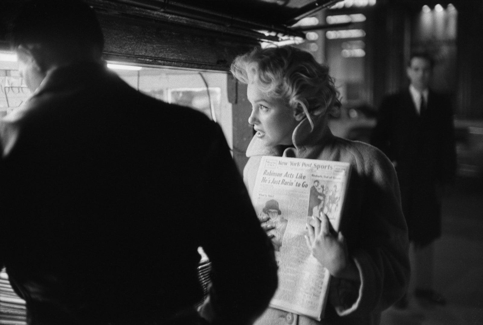 Black and white photograph of Marilyn Monroe holding a newspaper at a New York newsstand at night