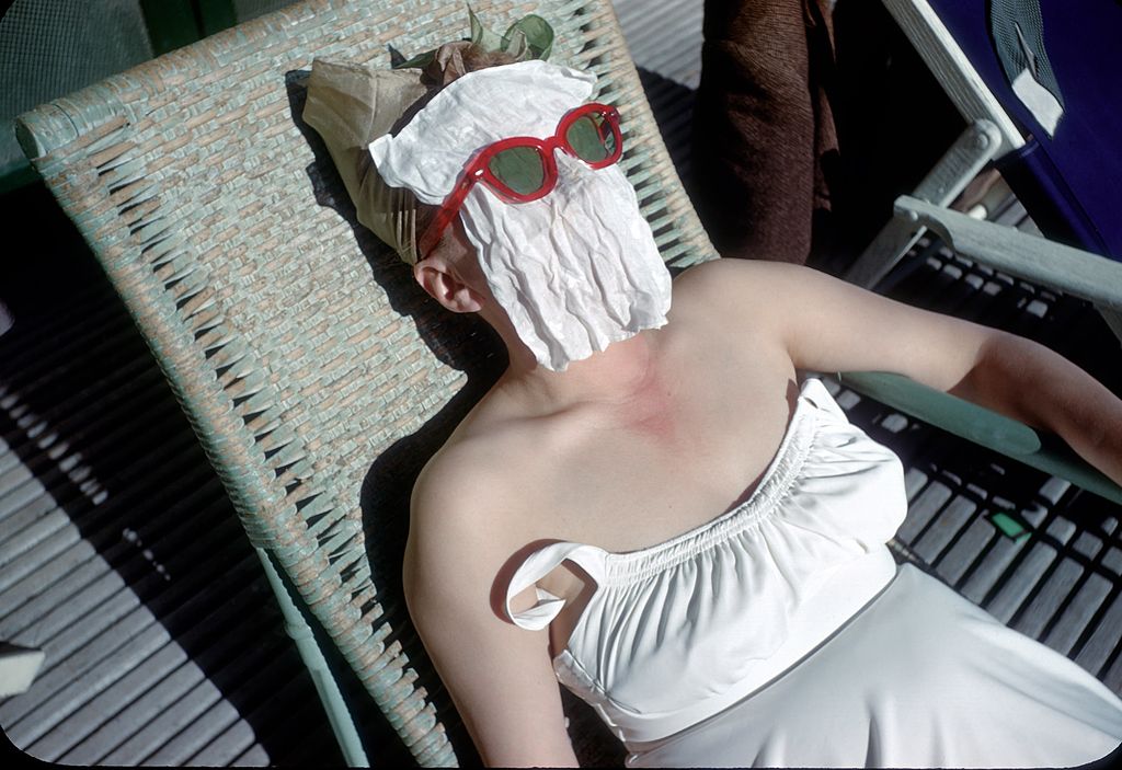 Colour photograph of a sunbather in a white strapless top lying on a lounger with a towel and red sunglasses over her face, 1949