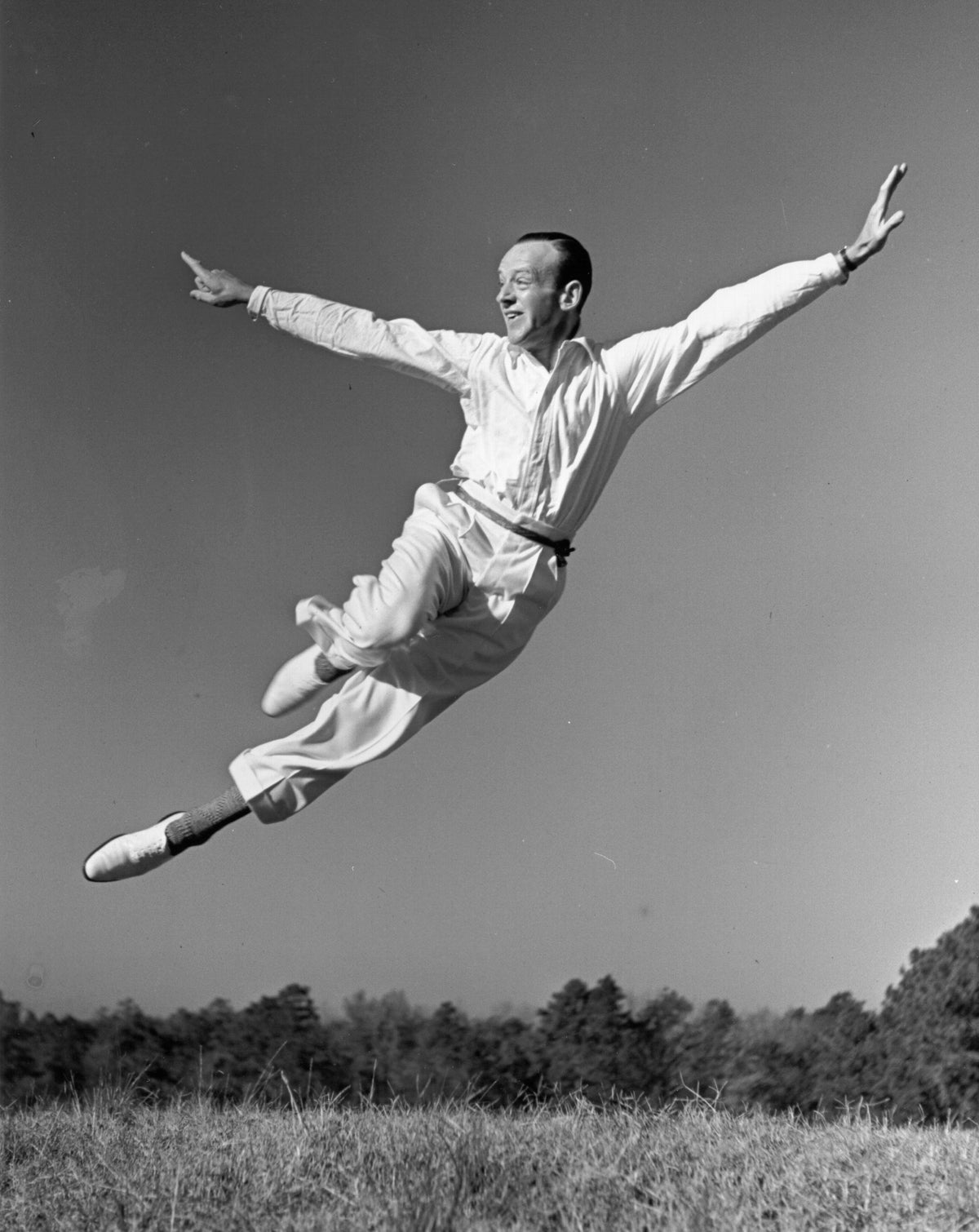 Black and white photograph of Fred Astaire leaping through the air with arms outstretched in an open field, circa 1950