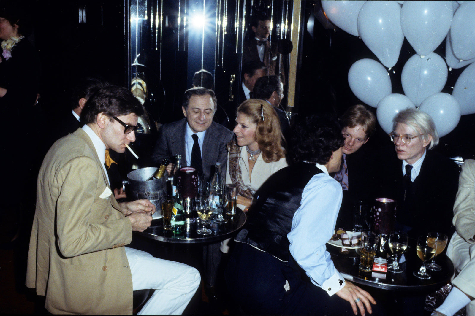 Colour photograph of Yves Saint Laurent, Pierre Berge and Andy Warhol seated at a table at a party at Le Palace, Paris, 1977