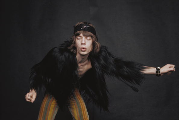 Colour portrait of Mick Jagger in a fur-trimmed jacket, arms outstretched, against a dark background, London, 1968