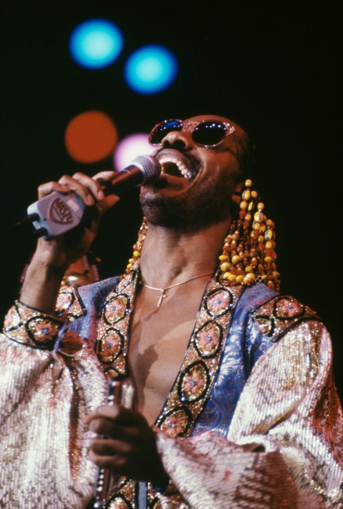 Colour photograph of Stevie Wonder singing into a microphone in sunglasses and a beaded embroidered top on stage at Wembley Arena, 1980