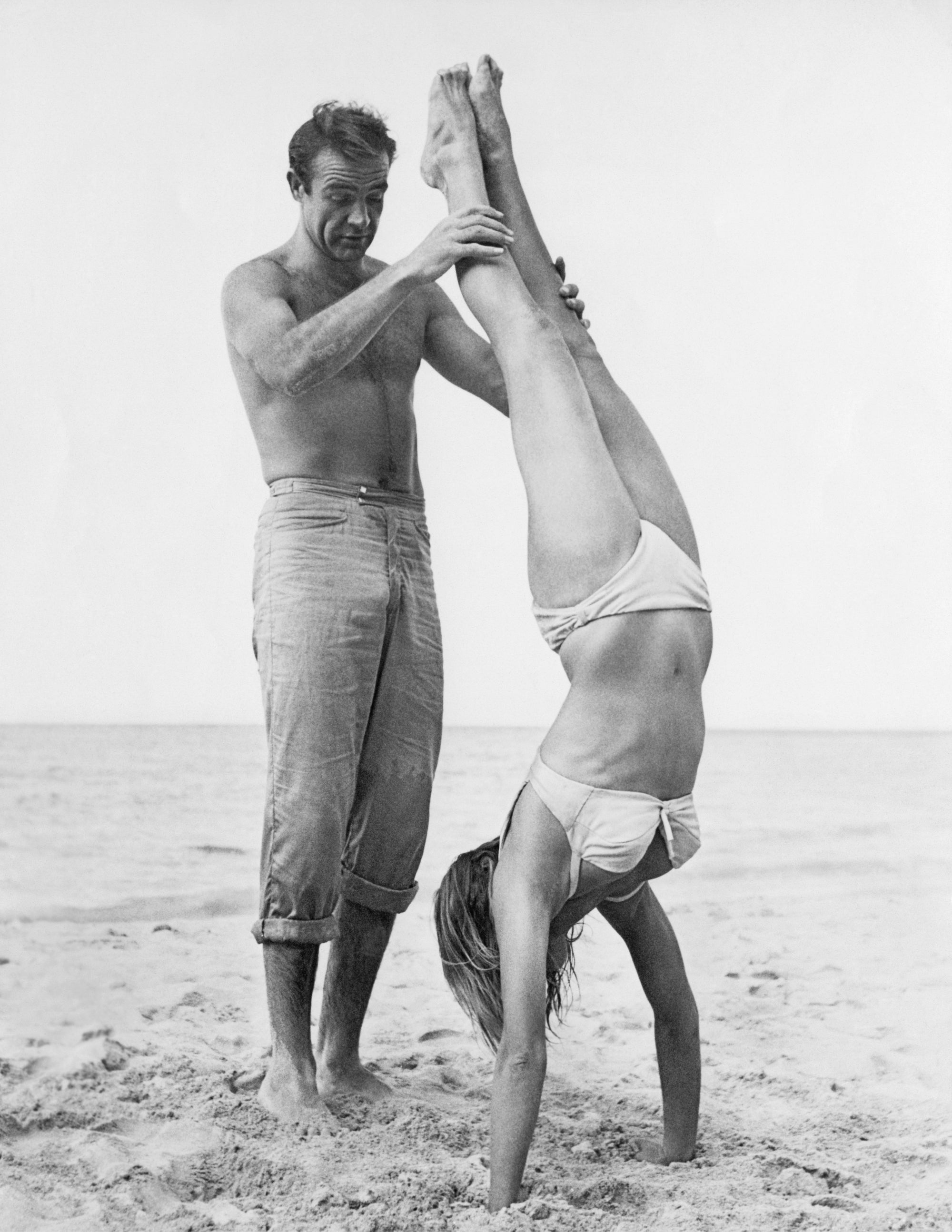 007 Catches Ursula by Hulton Archive/Getty Images, 1962. Shirtless Scottish actor Sean Connery holds Swiss actor Ursula Andress while she does a handstand on a beach in a white bikini on the set of the film, 'Dr.. Open Edition Resin Photographic Print. Portrait orientation. Available at Electric Gallery, London, UK.