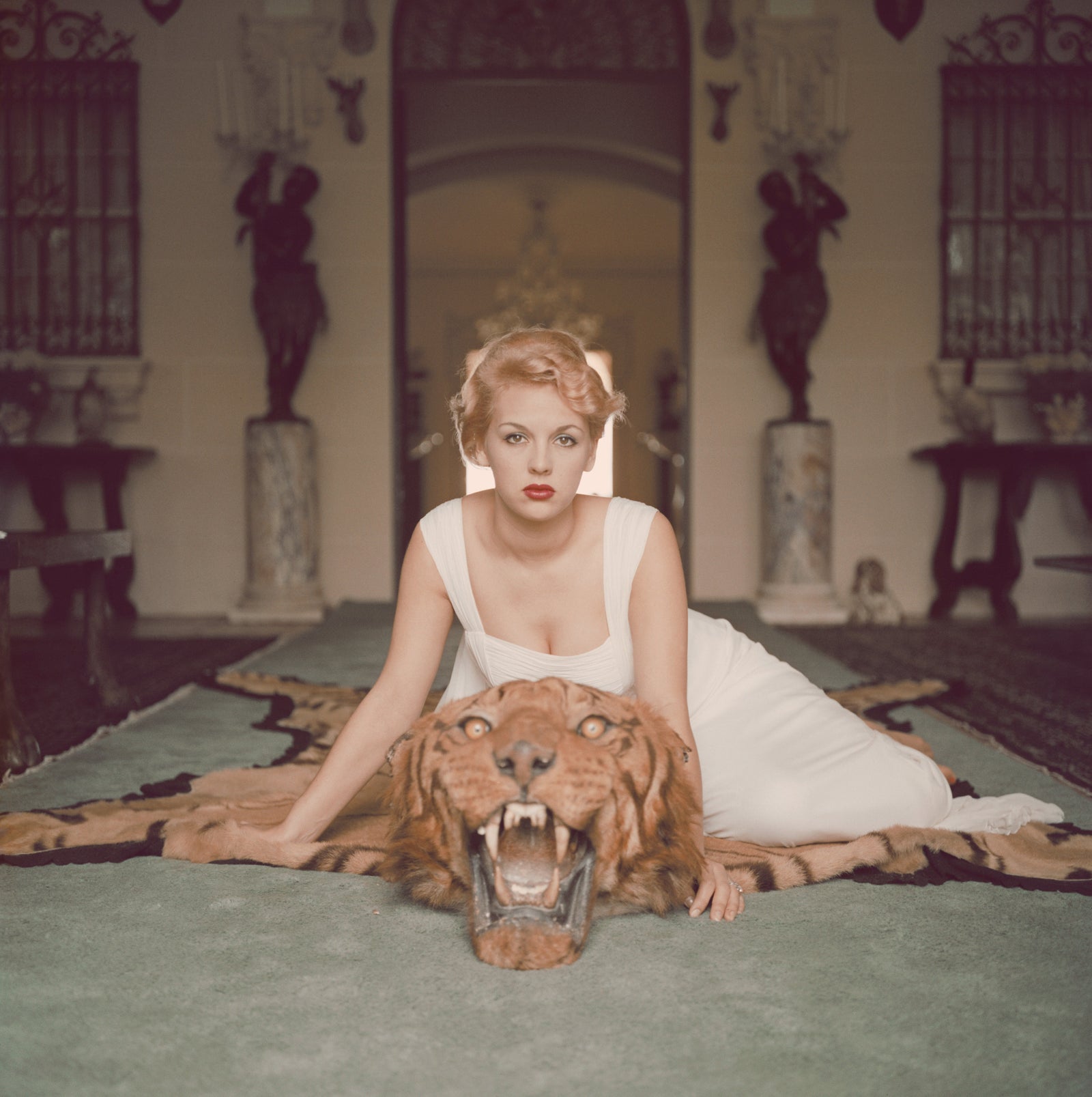 Beauty And The Beast by Slim Aarons/Getty Images. Lady Daphne Cameron on a tiger skin rug in the trophy room at socialite Laddie Sanford's home in Palm Beach, Florida. Open Edition C-Type Photographic Print. Square orientation. Available at Electric Gallery, London, UK.