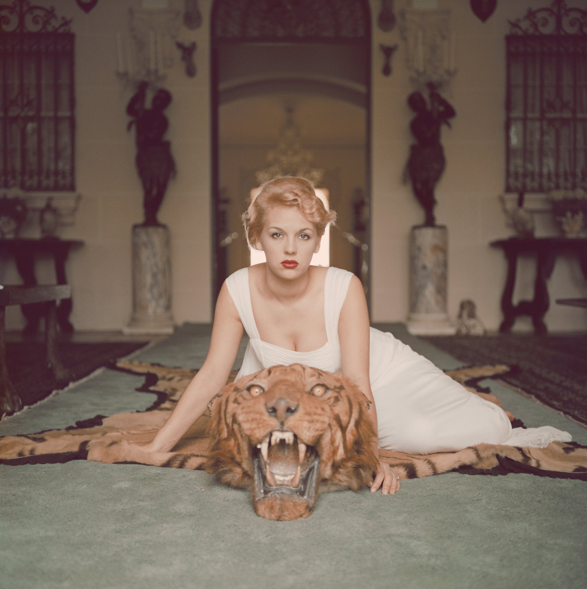 Beauty And The Beast by Slim Aarons/Getty Images. Lady Daphne Cameron on a tiger skin rug in the trophy room at socialite Laddie Sanford's home in Palm Beach, Florida. Open Edition C-Type Photographic Print. Square orientation. Available at Electric Gallery, London, UK.