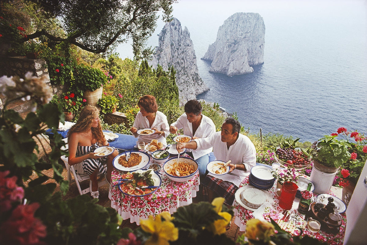 Dining Al Fresco On Capri by Slim Aarons/Getty Images, 1980. Italian artist and actress Domiziana Giordano, Italian author Francesca Sanvitale, Dino Trappetti and Umberto Terrelli dining al fresco on a terrace overlooking the waters off the coast of the island of Capri, Italy, in. Open Edition C-Type Photographic Print. Landscape orientation. Available at Electric Gallery, London, UK.