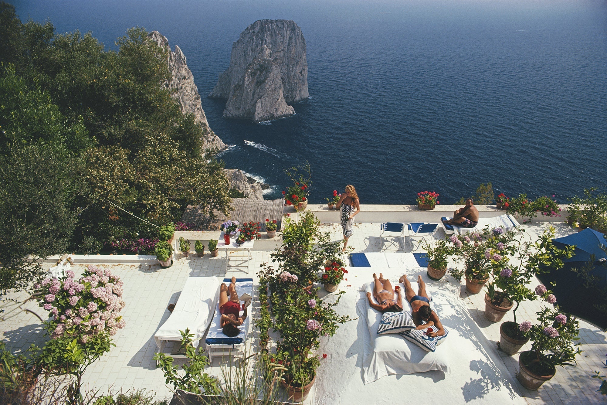 Il Canille by Slim Aarons/Getty Images, 1980. Sunbathers lounge on the white-painted terrace of Il Canille, built into the rocks of Pizzolungo overlooking the waters off the coast of the island of Capri, Italy, in. Open Edition C-Type Photographic Print. Landscape orientation. Available at Electric Gallery, London, UK.