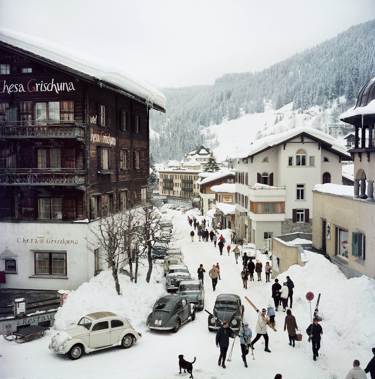 Klosters by Slim Aarons/Getty Images, 1963. Skiers pass by the Hotel Chesa Grischuna in Klosters. Open Edition C-Type Photographic Print. Square orientation. Available at Electric Gallery, London, UK.