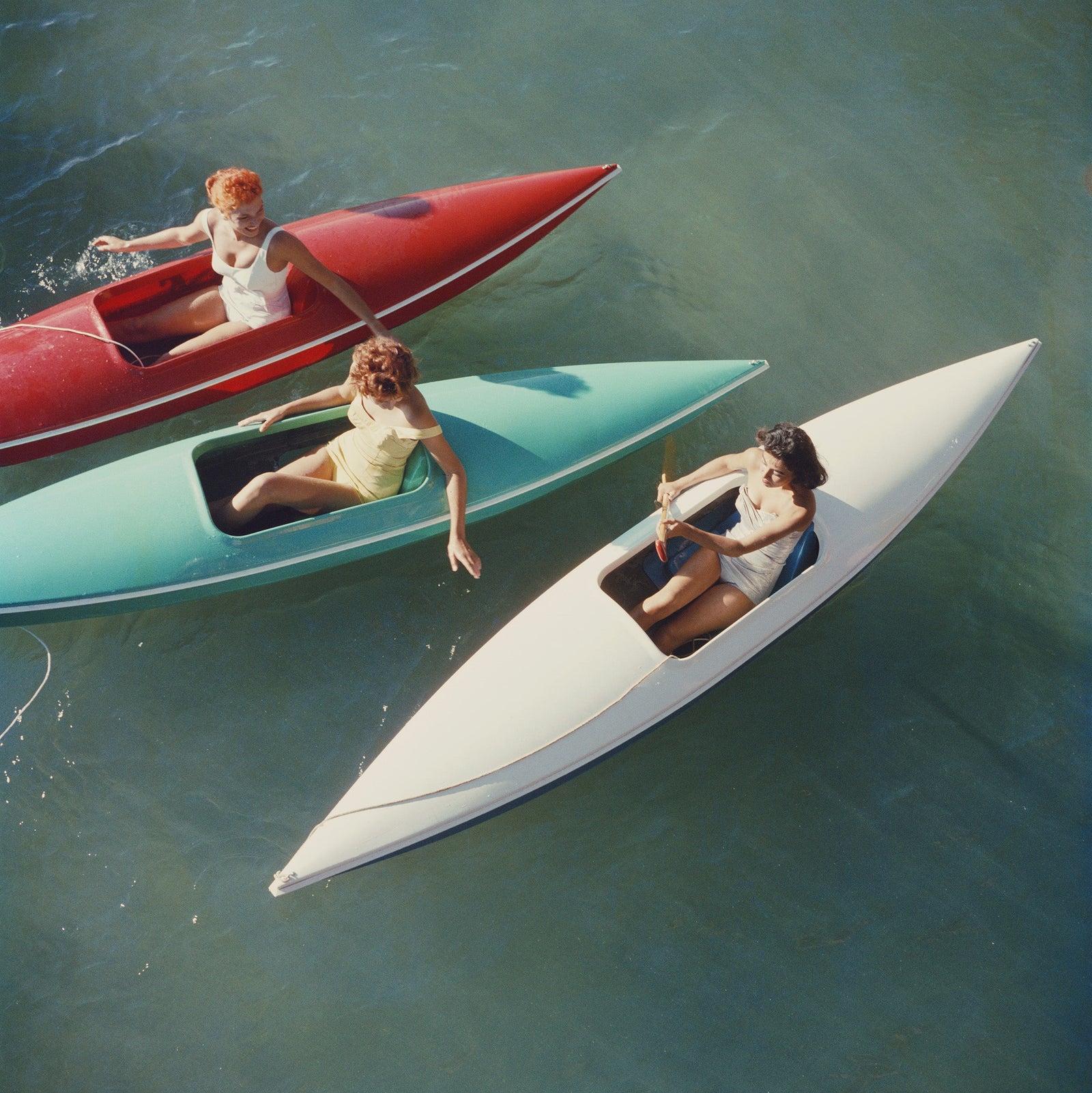Lake Tahoe Trip by Slim Aarons/Getty Images, 1959. Young women canoeing at Zephyr Cove on the Nevada side of Lake Tahoe, USA. Open Edition C-Type Photographic Print. Square orientation. Available at Electric Gallery, London, UK.