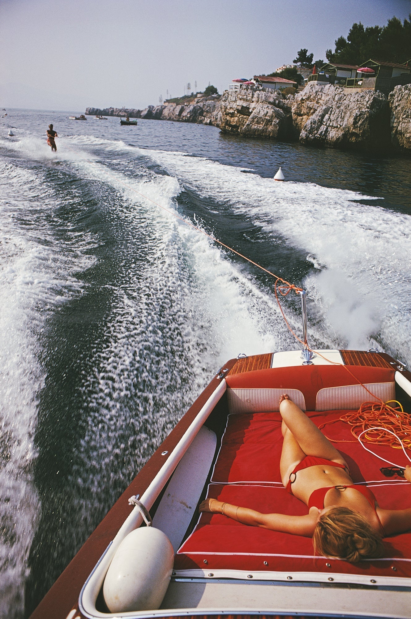 Leisure In Antibes by Slim Aarons/Getty Images, 1969. A woman sunbathing in a motorboat as it tows a waterskiier, in the sea off the Hotel du Cap-Eden-Roc in Antibes on the French Riviera. Open Edition C-Type Photographic Print. Portrait orientation. Available at Electric Gallery, London, UK.