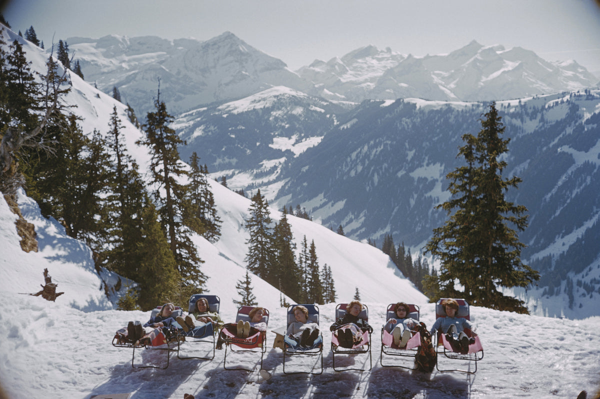 Lounging In Gstaad by Slim Aarons/Getty Images, 1961. Holidaymakers in sun loungers on the slopes at Gstaad, Switzerland
 . Open Edition C-Type Photographic Print. Landscape orientation. Available at Electric Gallery, London, UK.