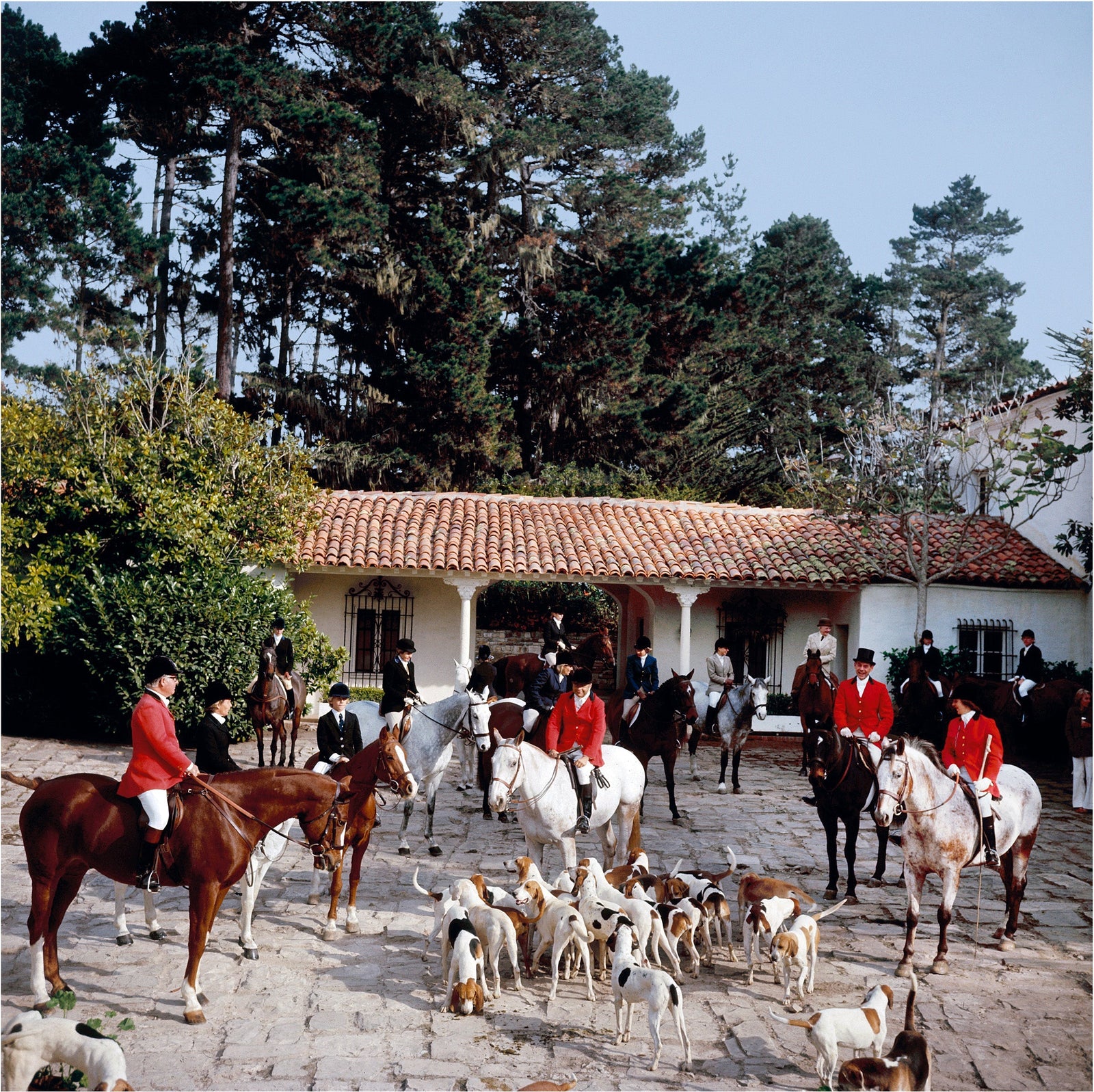 Pebble Beach Hunt by Slim Aarons/Getty Images, 1976. Riders gather in the courtyard of Richard Collins' House for the stirrup cup libation, before the start of the Pebble Beach Hunt of English and American foxhounds. Open Edition C-Type Photographic Print.. Square orientation. Available at Electric Gallery, London, UK.