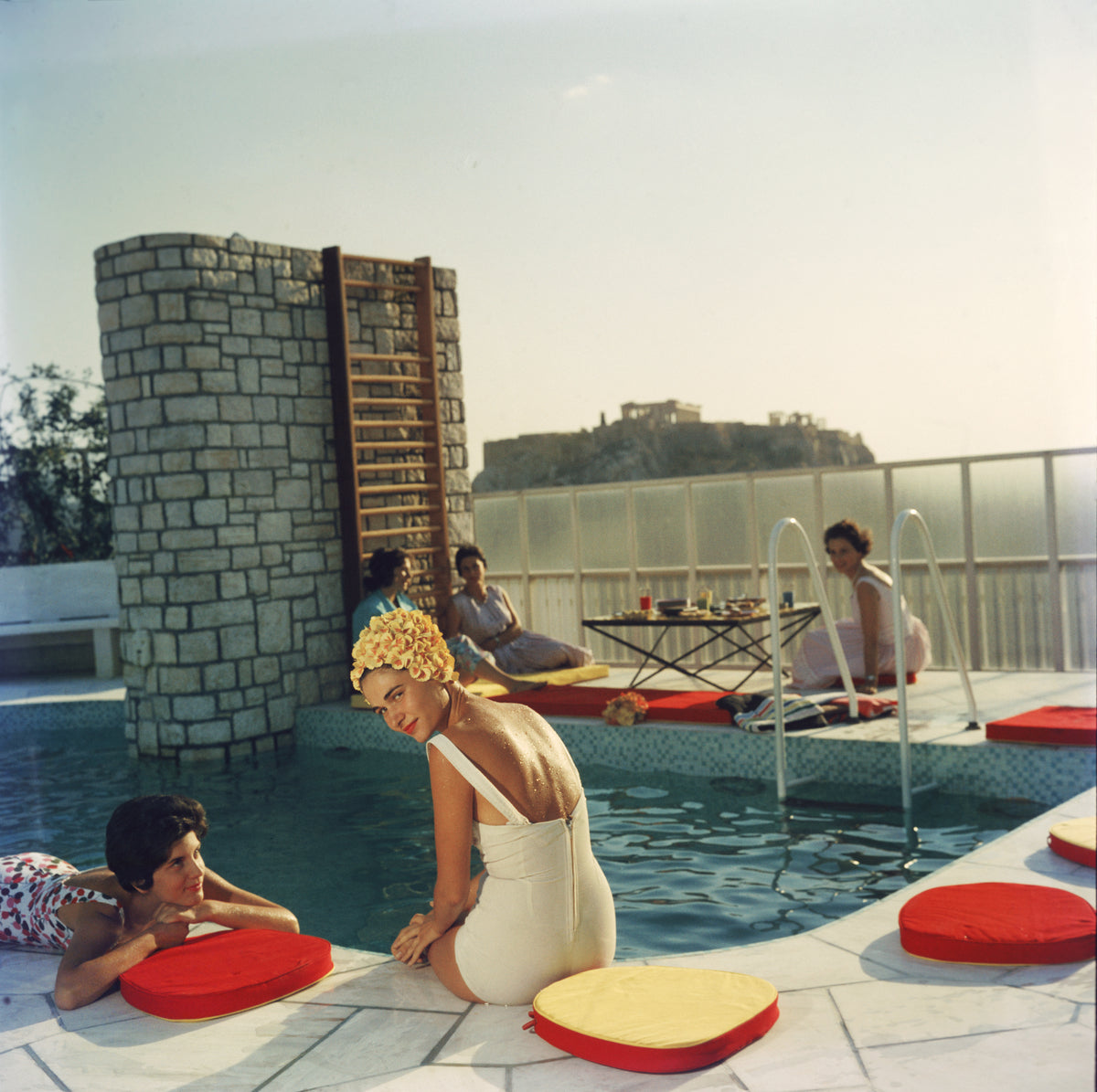 Penthouse Pool by Slim Aarons/Getty Images, 1961. Young women by the Canellopoulos penthouse pool, Athens. Open Edition C-Type Photographic Print. Square orientation. Available at Electric Gallery, London, UK.